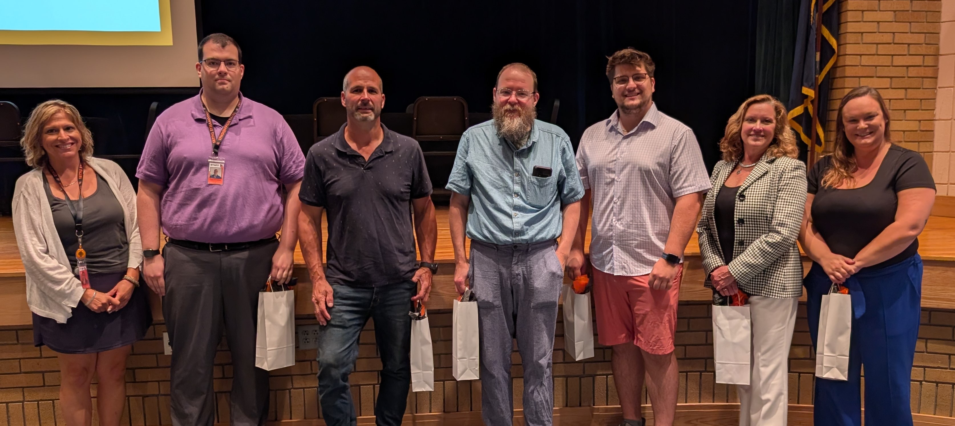 A group of six adults stand in a row on a wooden stage, each holding a small white gift bag.