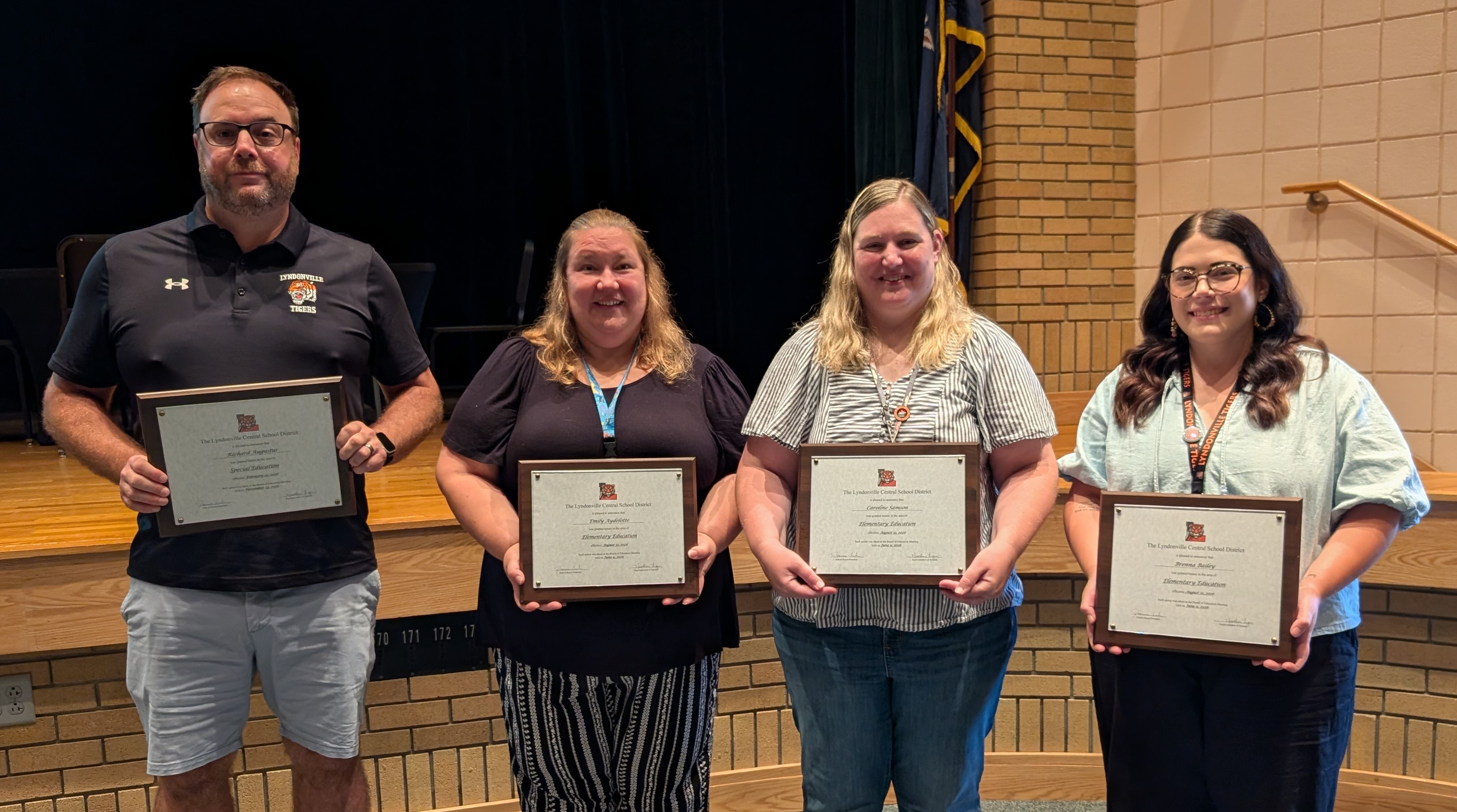 Four adults stand proudly on a stage in front of a brick wall, each holding a framed award plaque from the Lyndonville Central School District.