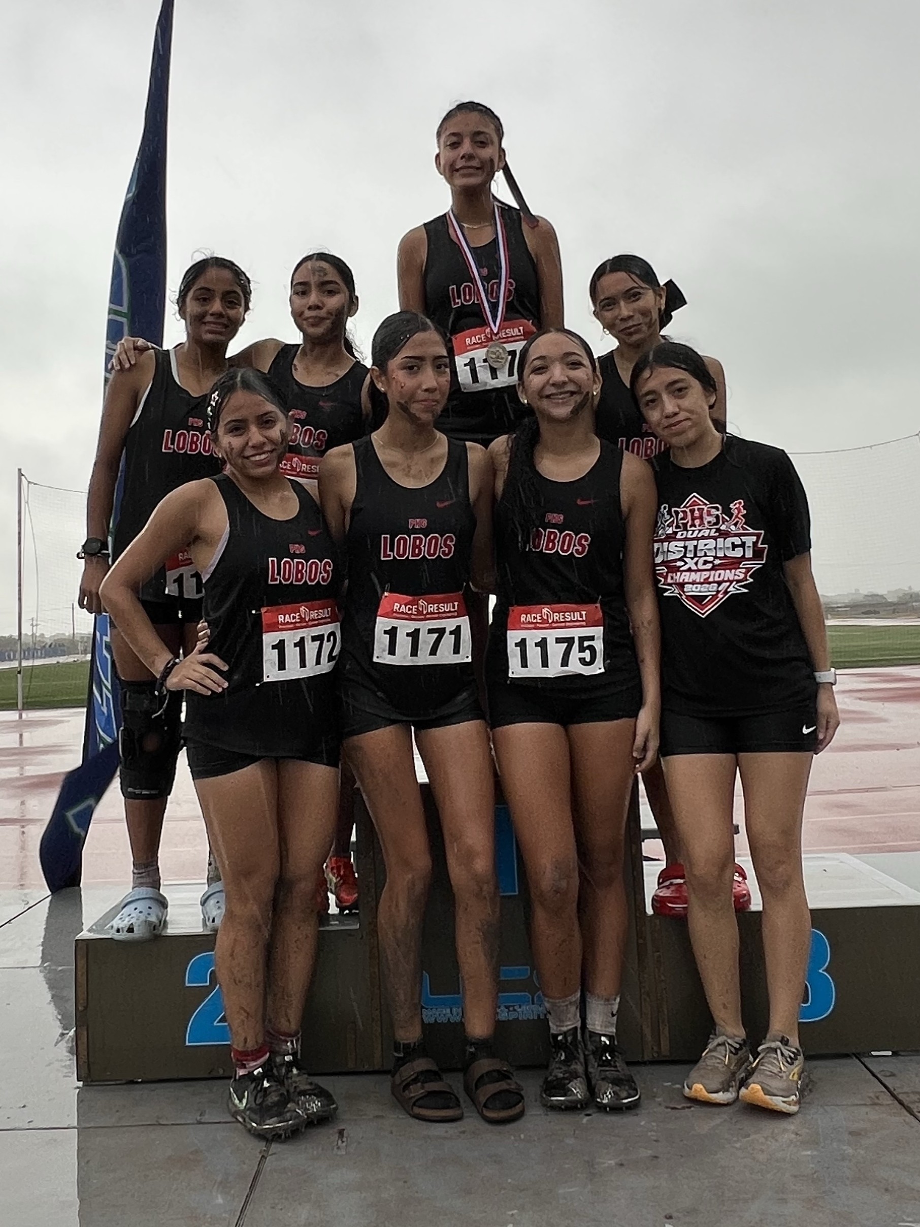 Seven female athletes in matching black Lobos uniforms stand proudly on a victory podium after a muddy race.