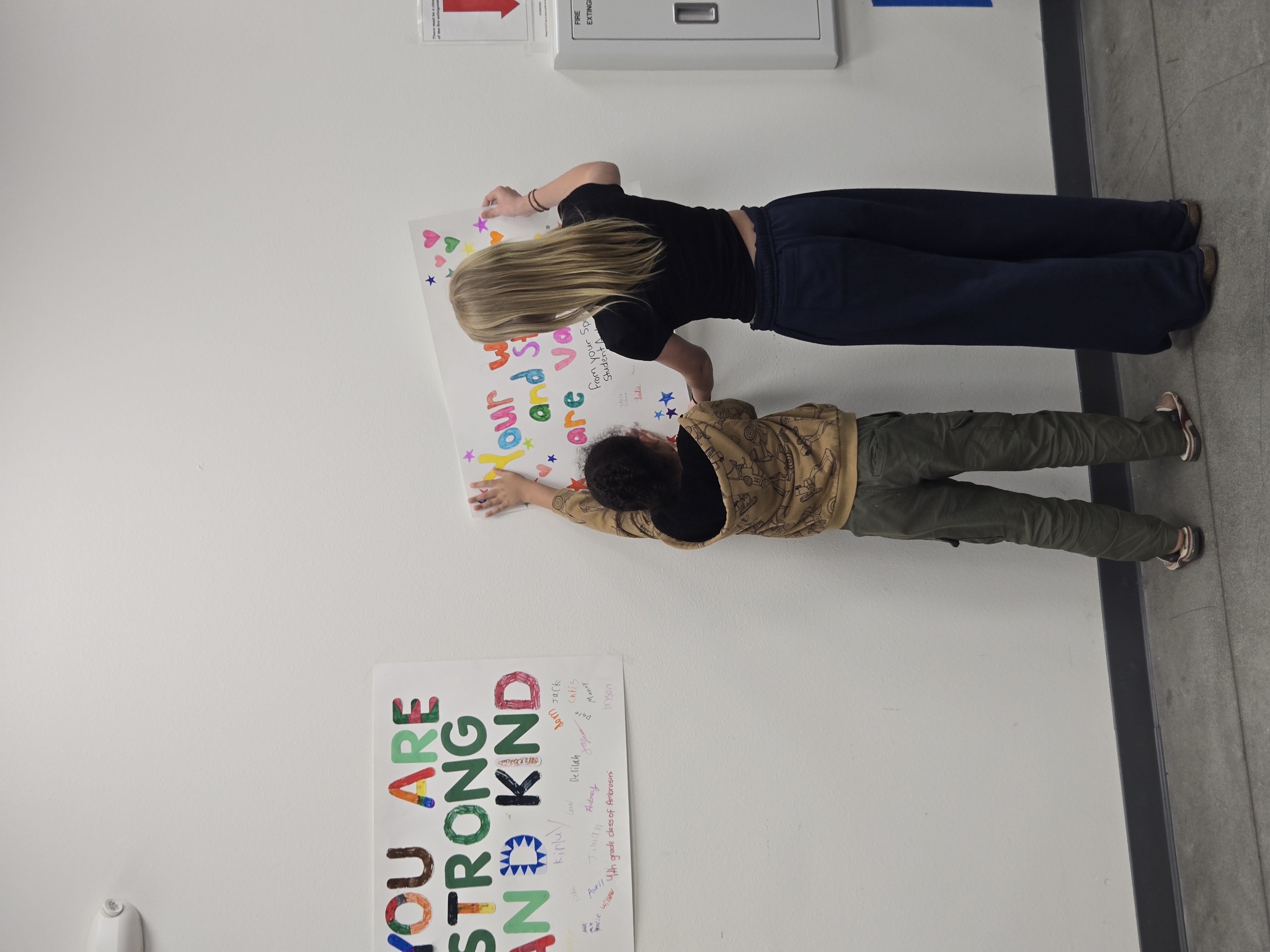 Two children are placing colorful stars and hearts on a poster that reads 'YOU ARE STRONG AND KIND'.