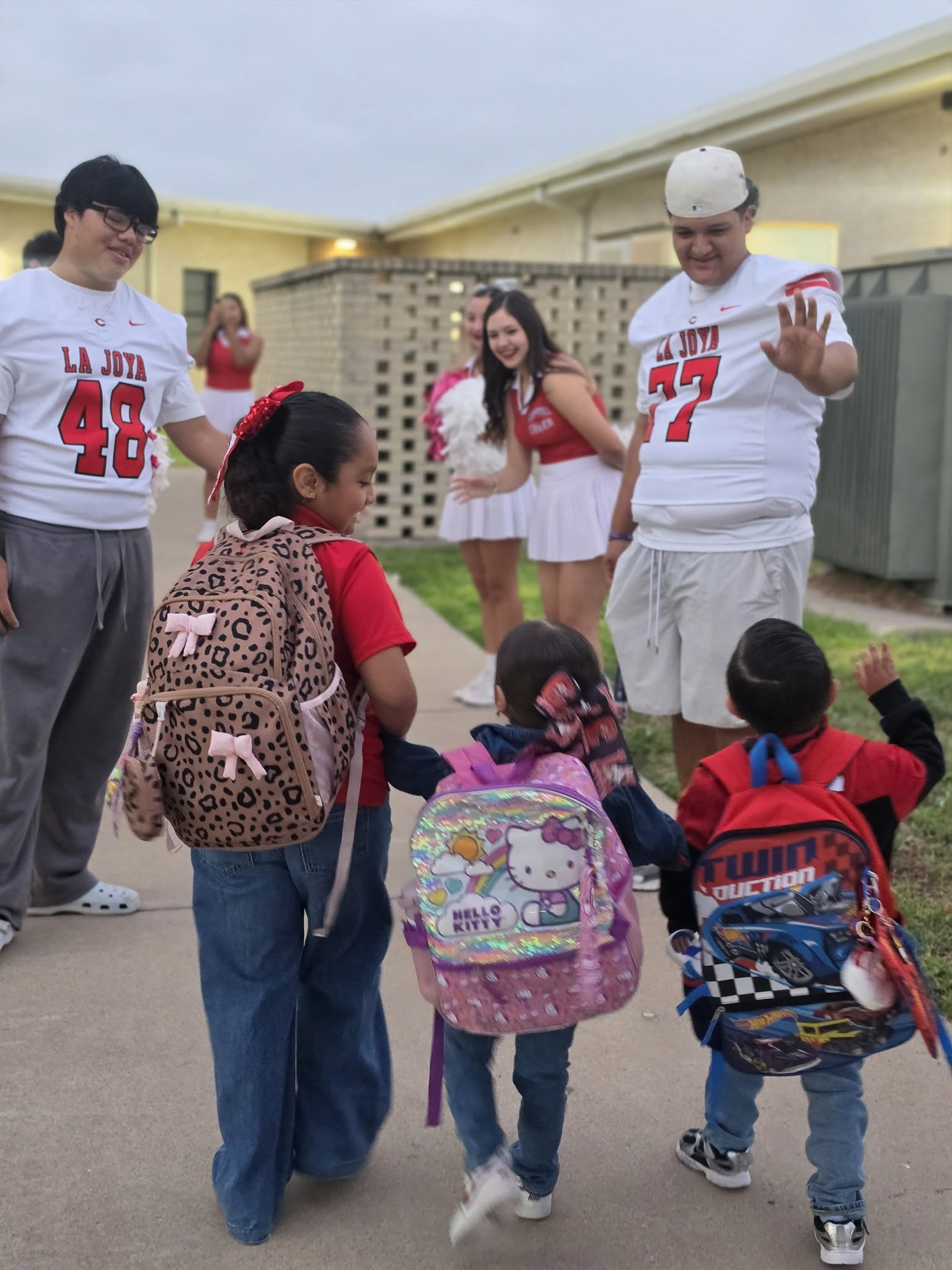 Three children with colorful backpacks are greeted by football players and cheerleaders.