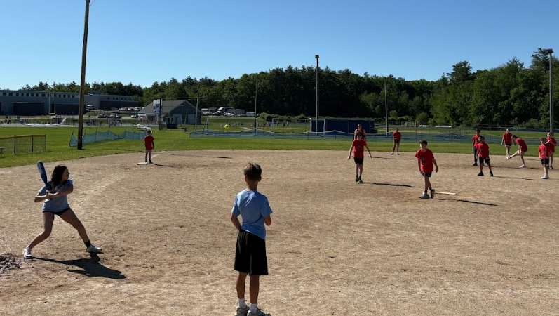 A young person swings a baseball bat on a dirt field with other players in the background.