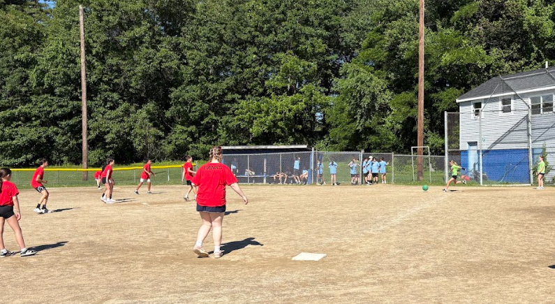 Children in red and blue uniforms play a game of kickball on a sunny day.