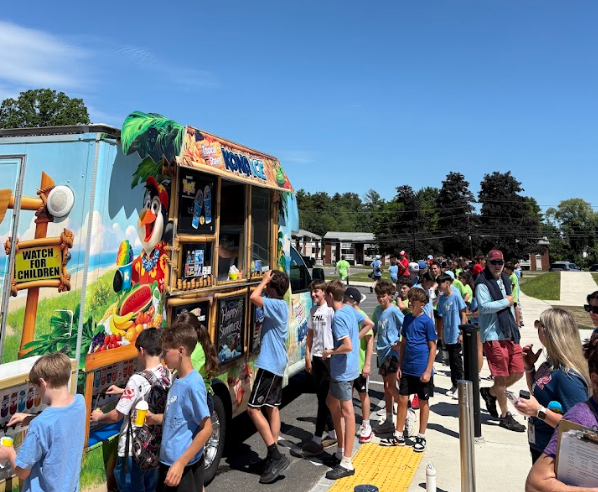 A line of children and adults wait to order from a colorful Kona Ice truck on a sunny day.