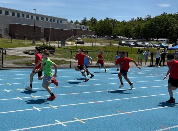 Children race on a bright blue track under a clear sky, with a school building in the background.