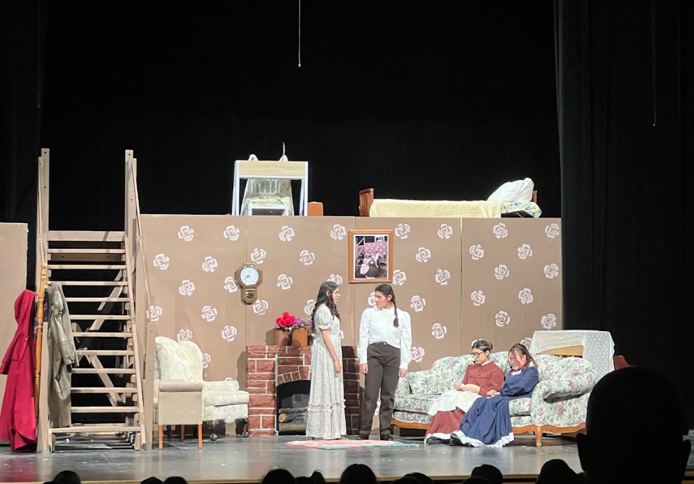 A theatrical stage set depicts a room with a fireplace, sofa, and beds, featuring actors in period costumes.