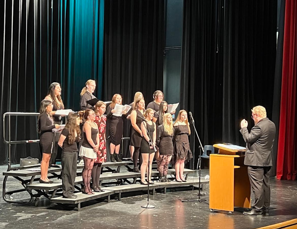 A choir of young people stands on risers on a stage, singing from sheet music.