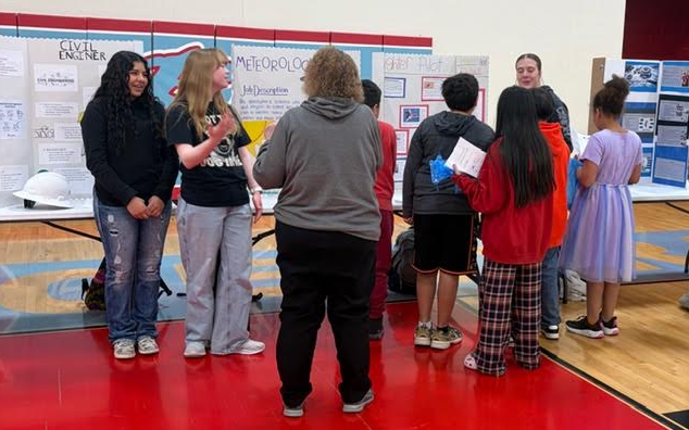 Students stand at a science fair, presenting projects on display boards.