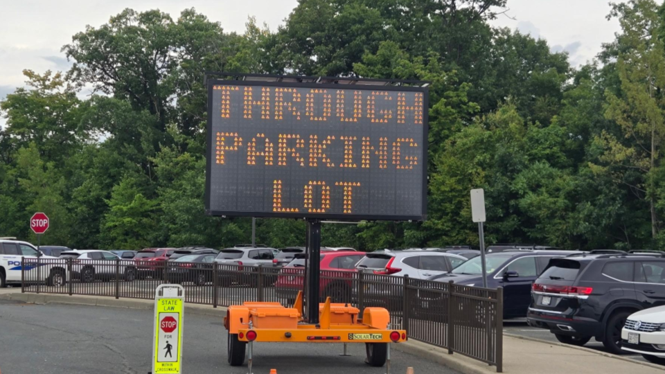 A digital portable message sign stands in front of a parking lot, displaying the text "THROUGH PARKING LOT".