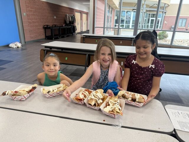 Three young girls smile while sitting behind a cafeteria table filled with paper boats containing rolled food topped with white sauce.