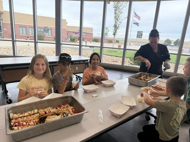 A group of children gather around a long table indoors, smiling as they prepare to eat rolled crepes from a catering pan.