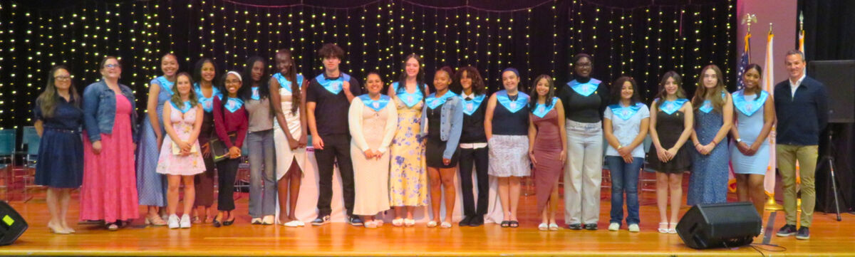 A diverse group of graduates in light blue sashes stand on a stage with a sparkling backdrop.