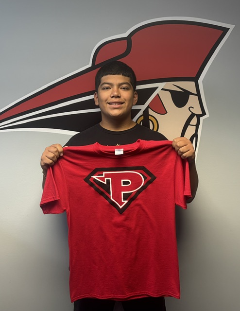 A young person stands smiling, holding up a red t-shirt with a white 'P' inside a diamond logo.