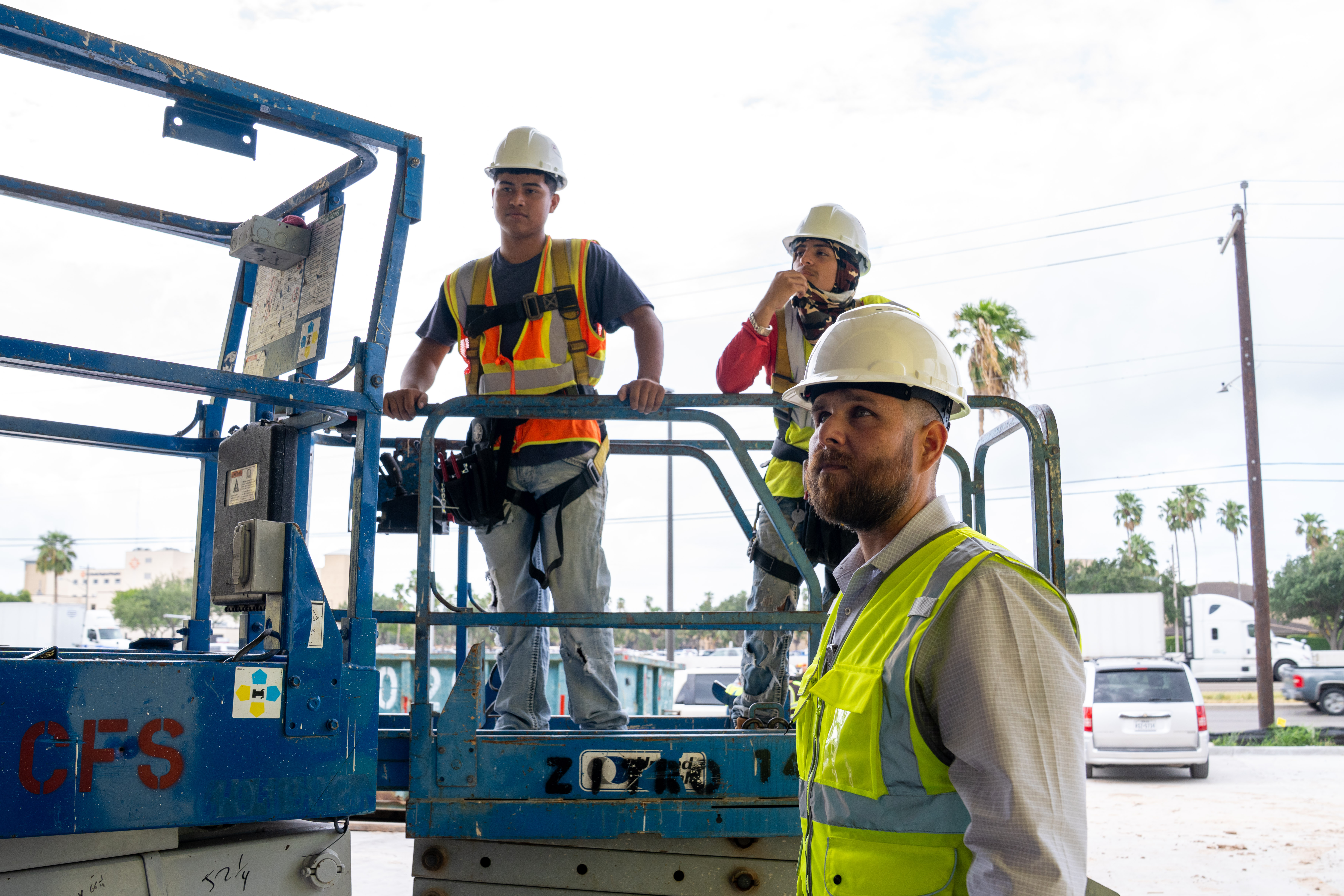 Three construction workers in hard hats and safety vests stand on a blue scissor lift.