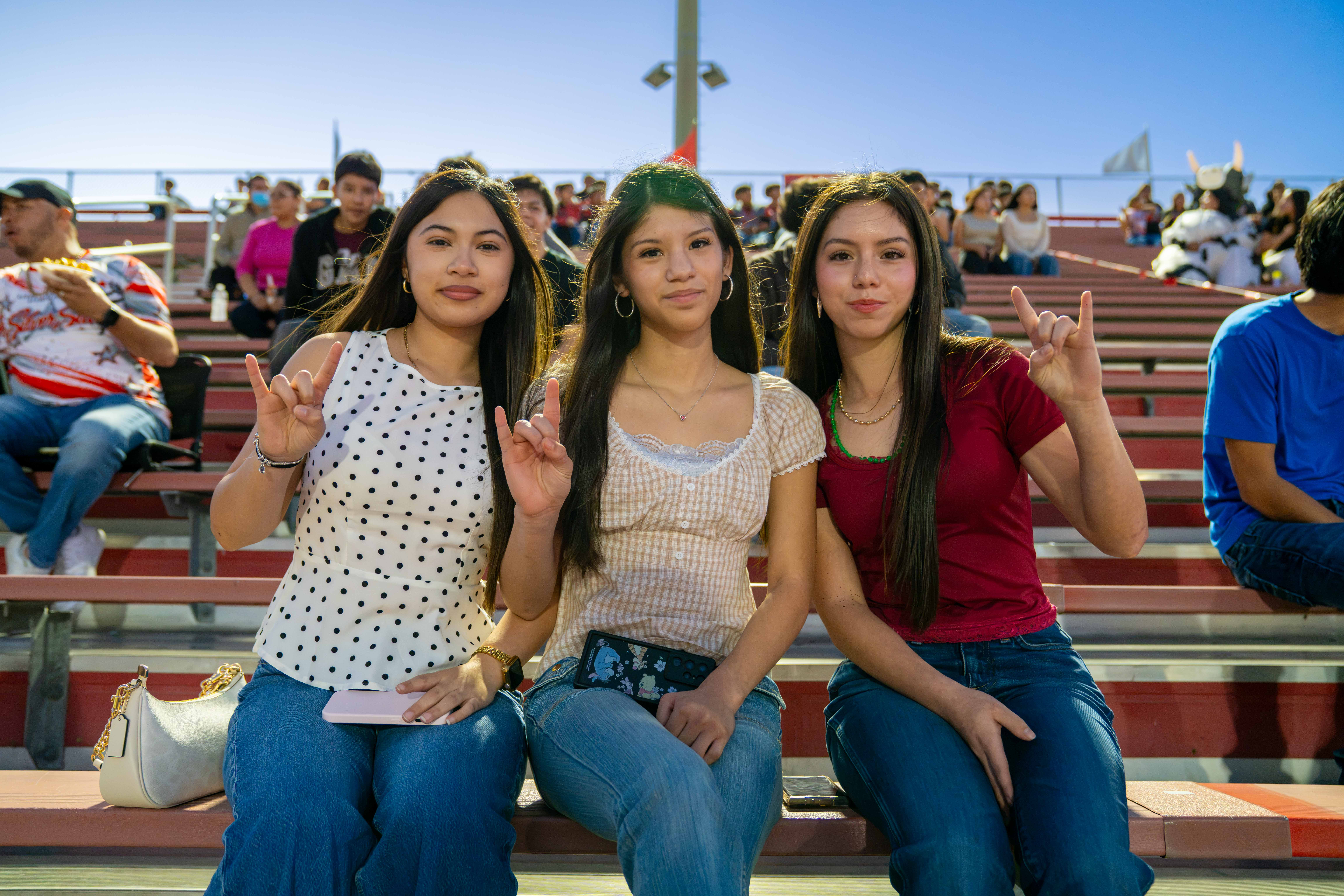 Three young women sit together in the stands, flashing rock-and-roll hand signs under a bright blue sky.