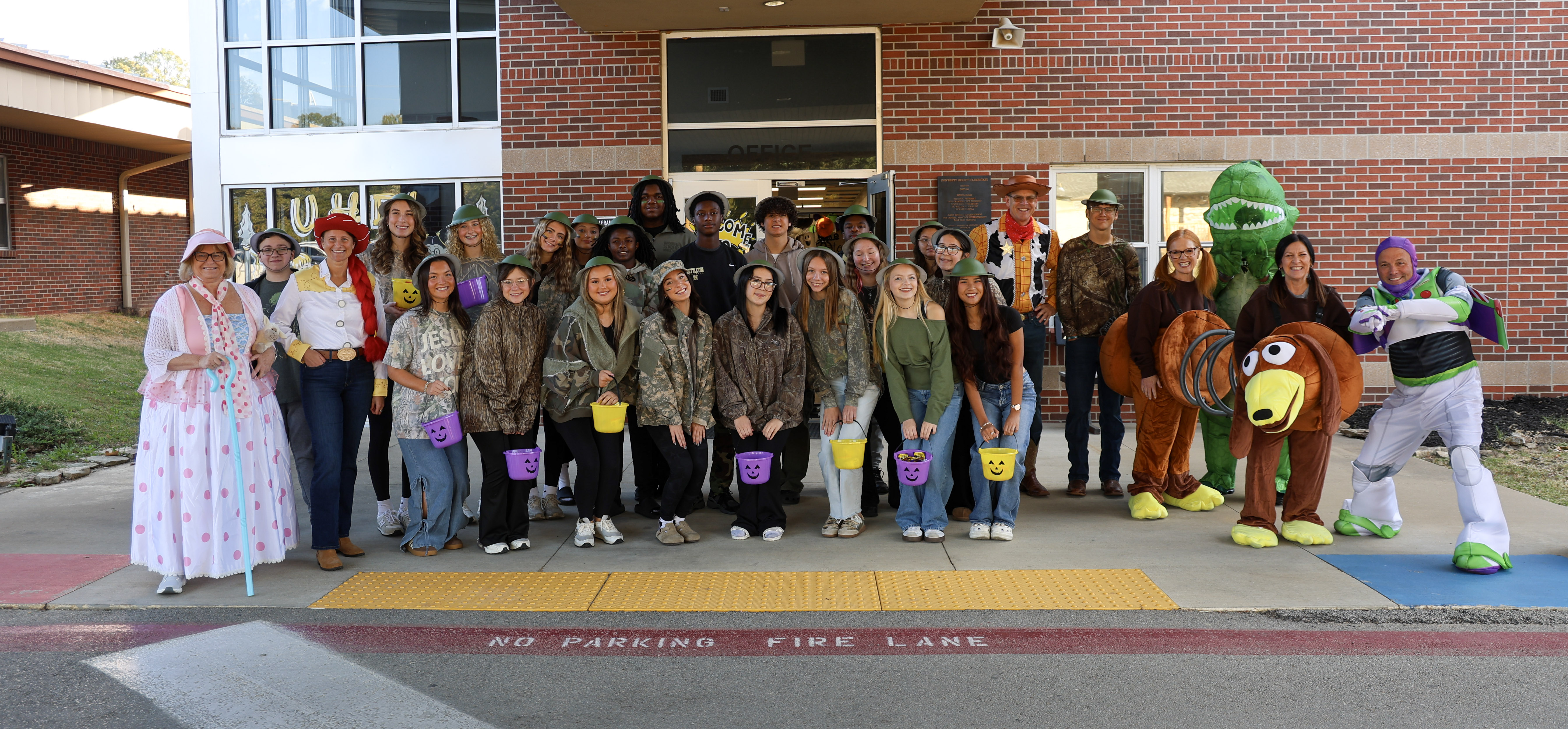A large group of people poses in front of a brick building wearing various costumes, including Toy Story characters and camouflage outfits with toy buckets.