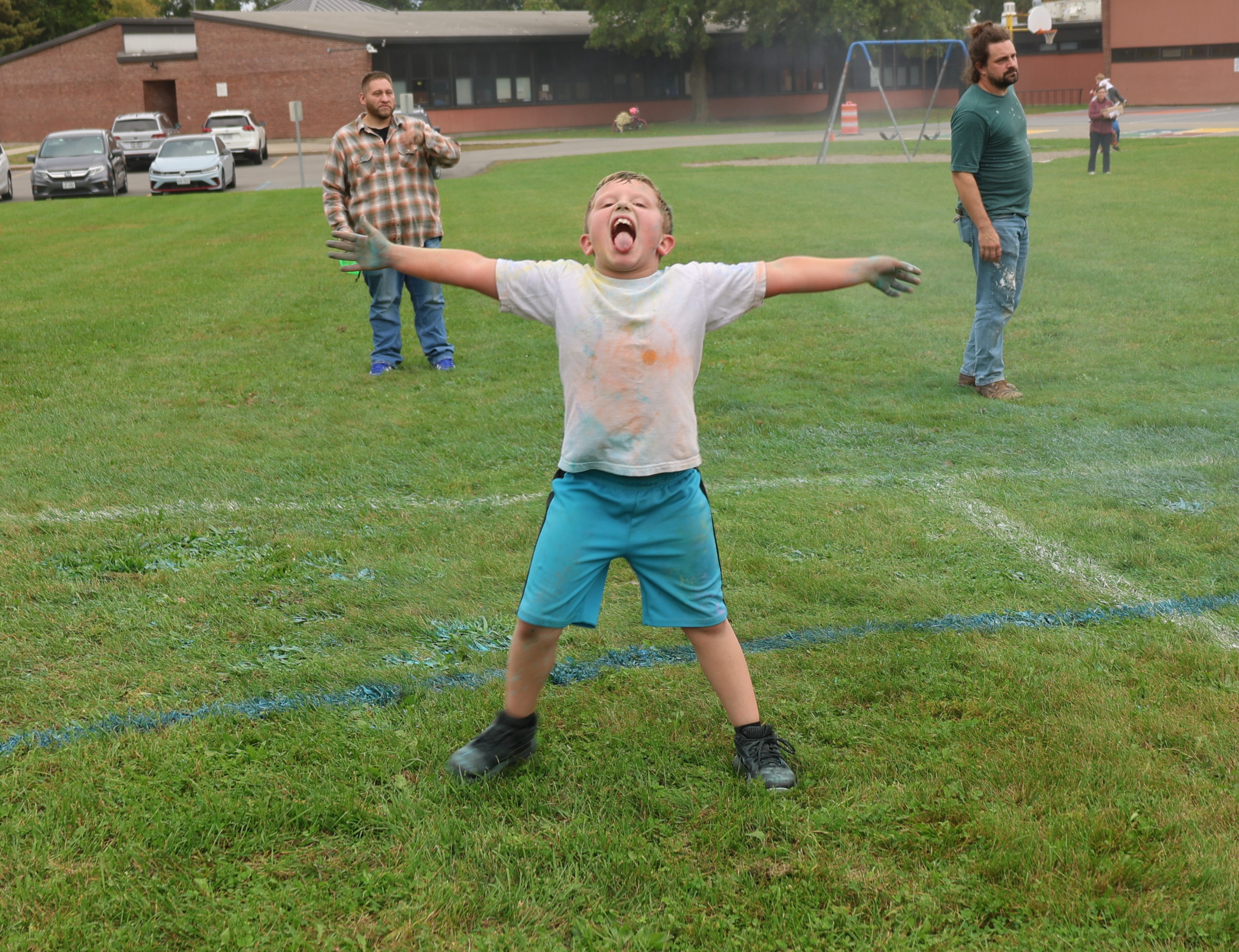 A child stands on a grassy field with arms outstretched and tongue out, wearing a color-splattered shirt and blue shorts.
