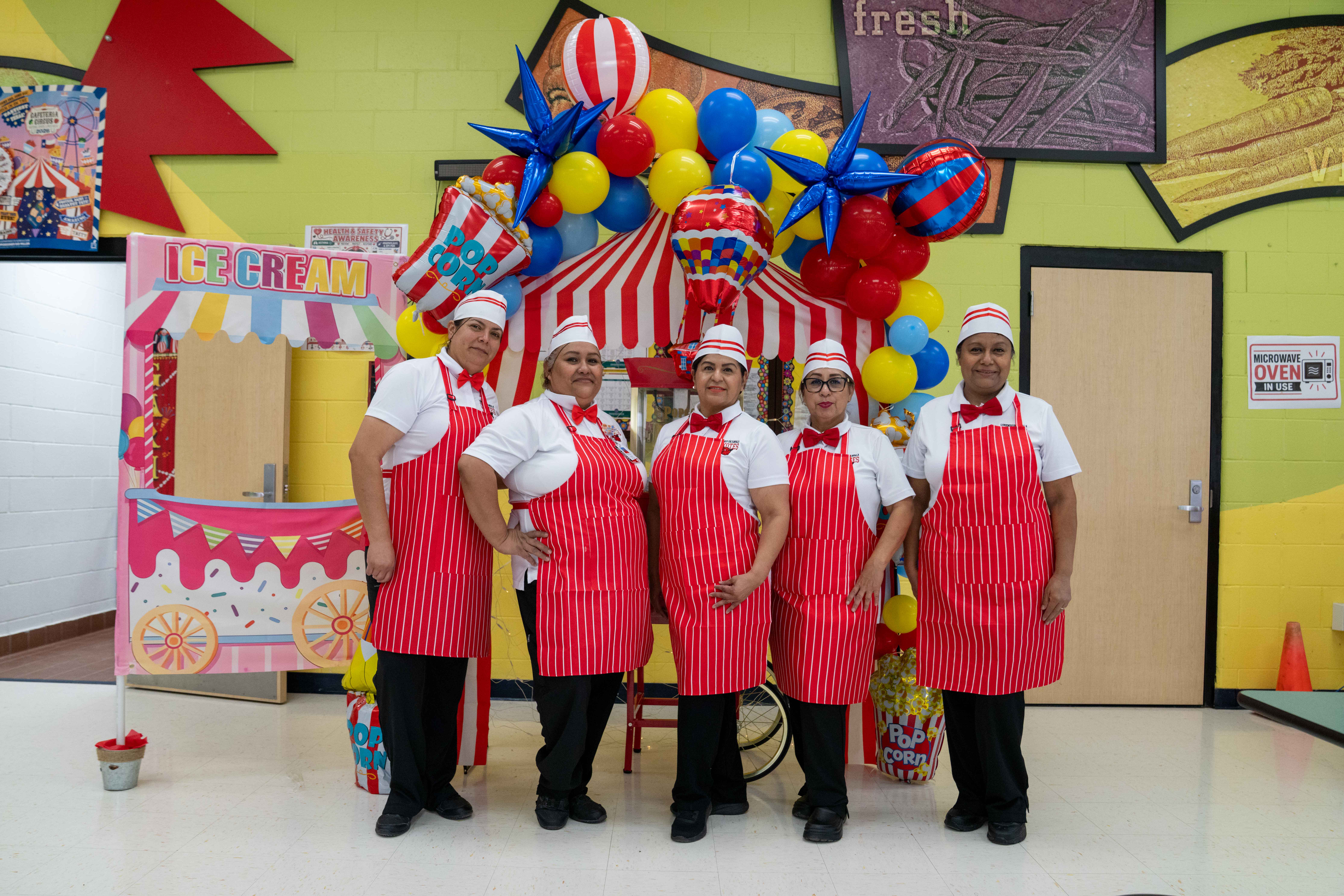 Five food service workers in matching red and white striped aprons and hats stand smiling in front of a carnival-themed booth.
