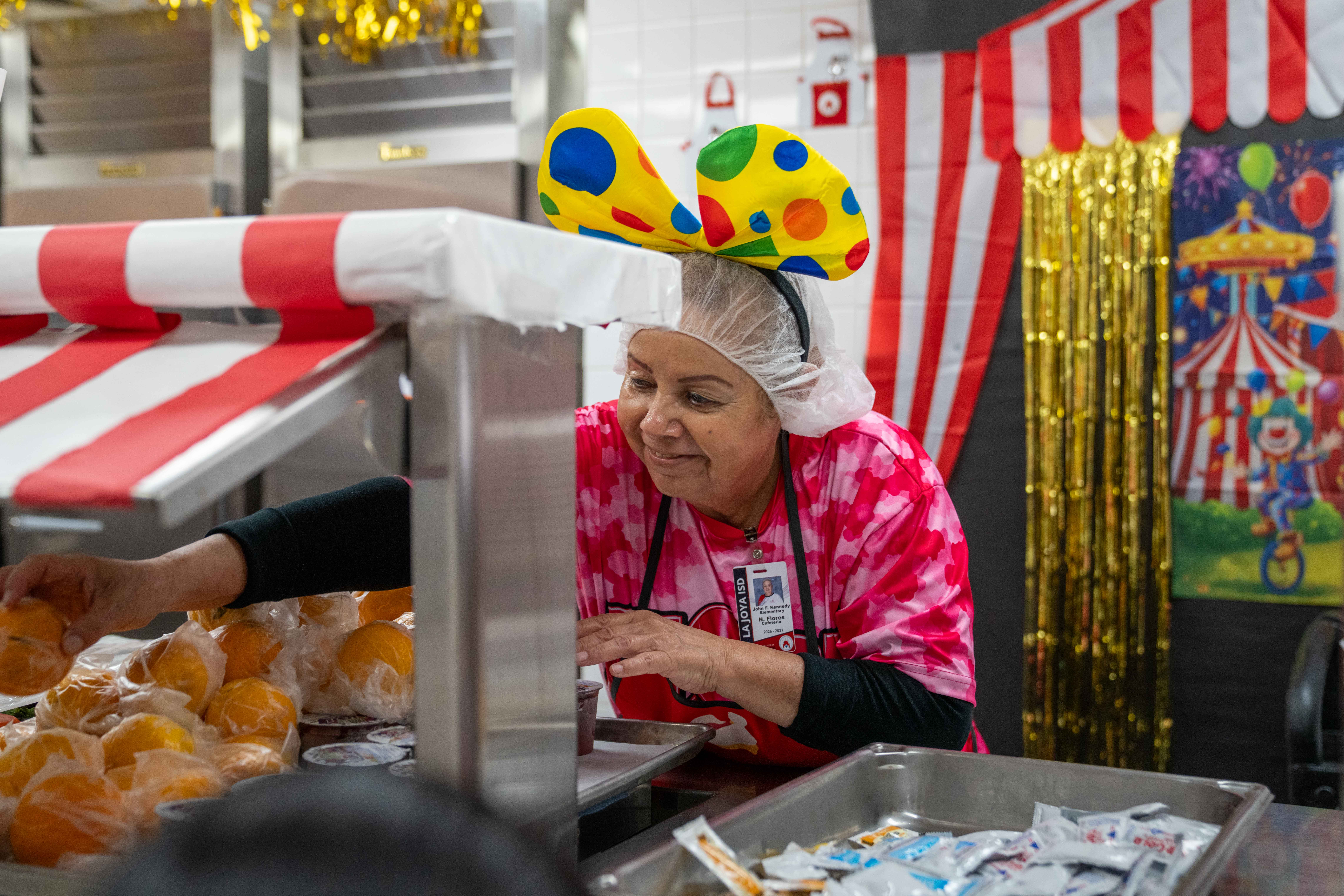 A cafeteria worker wearing a colorful polka dot headband and hairnet serves food from behind a carnival-themed counter.