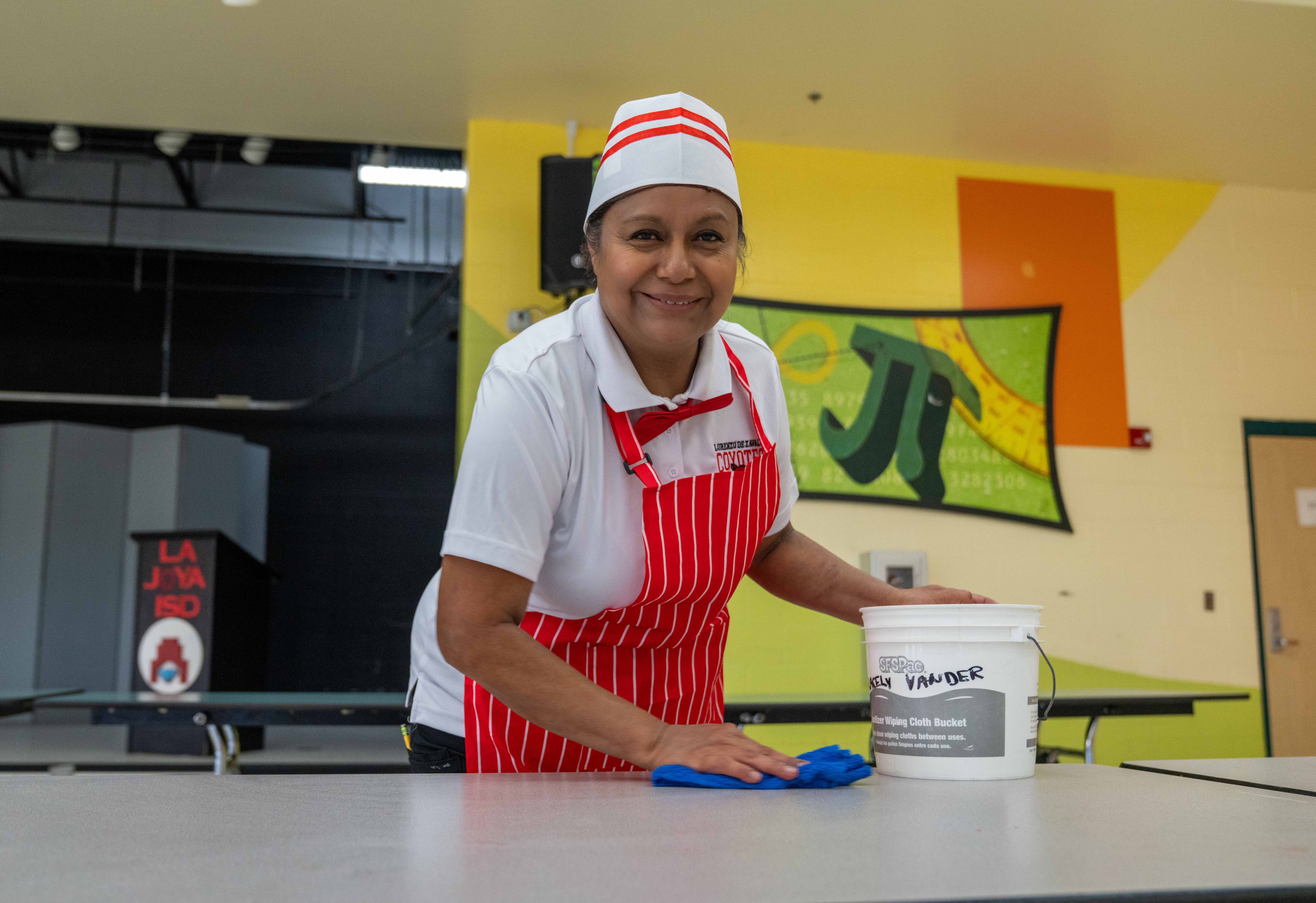 A smiling food service worker in a red-and-white striped apron and paper hat wipes down a cafeteria table.