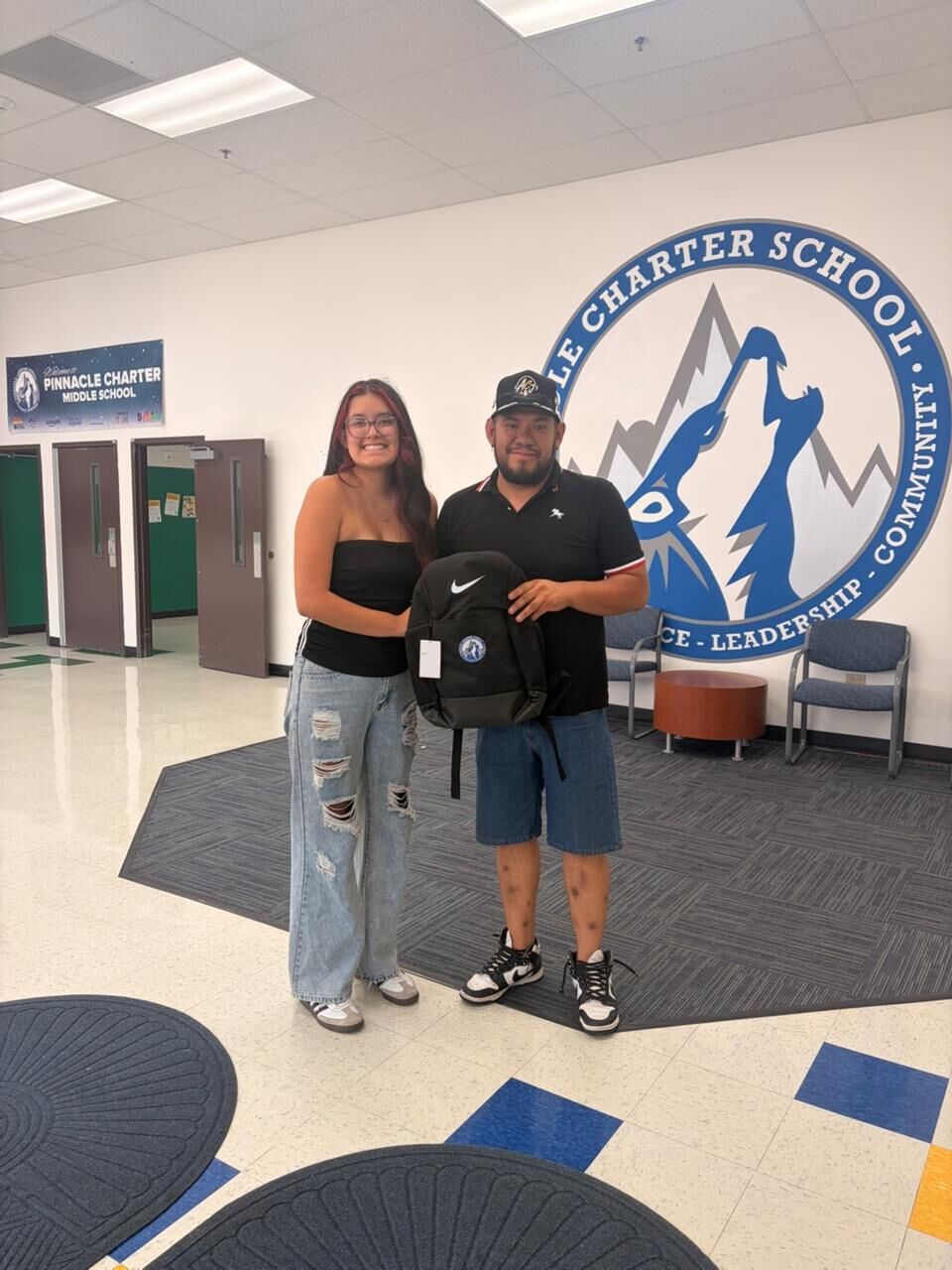 Two individuals pose for a photo while holding a black Nike backpack in the Pinnacle Charter School lobby, with the school's Timberwolf logo displayed on the wall behind them.