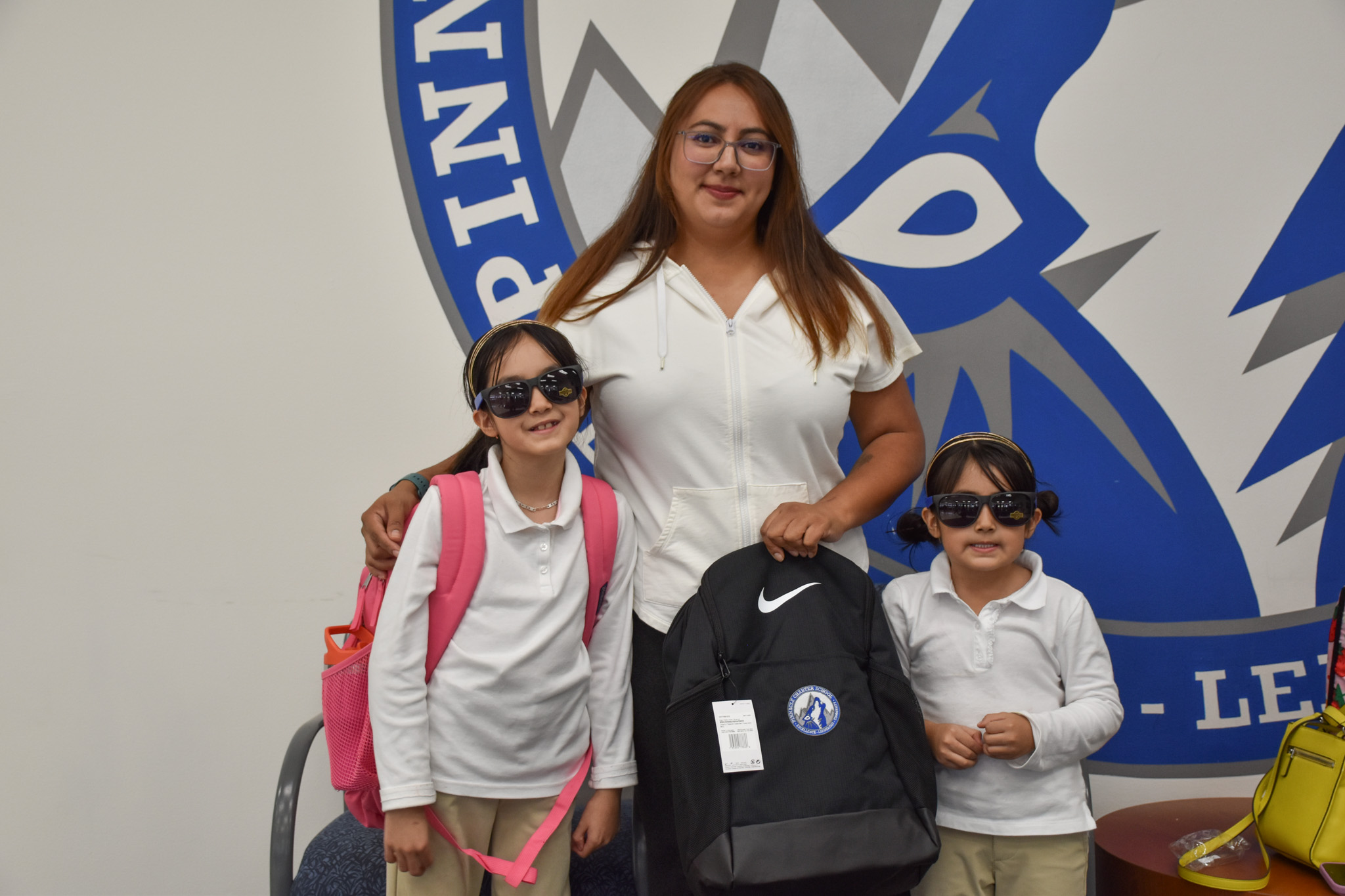 A woman stands with two young students wearing sunglasses and backpacks, posing in front of the Pinnacle Charter School Timberwolf logo while holding a black Nike backpack.