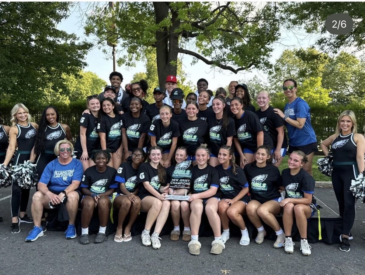 A diverse group of young women and coaches in black Eagles-themed shirts pose for a photo, holding a "New Jersey Champions" trophy.