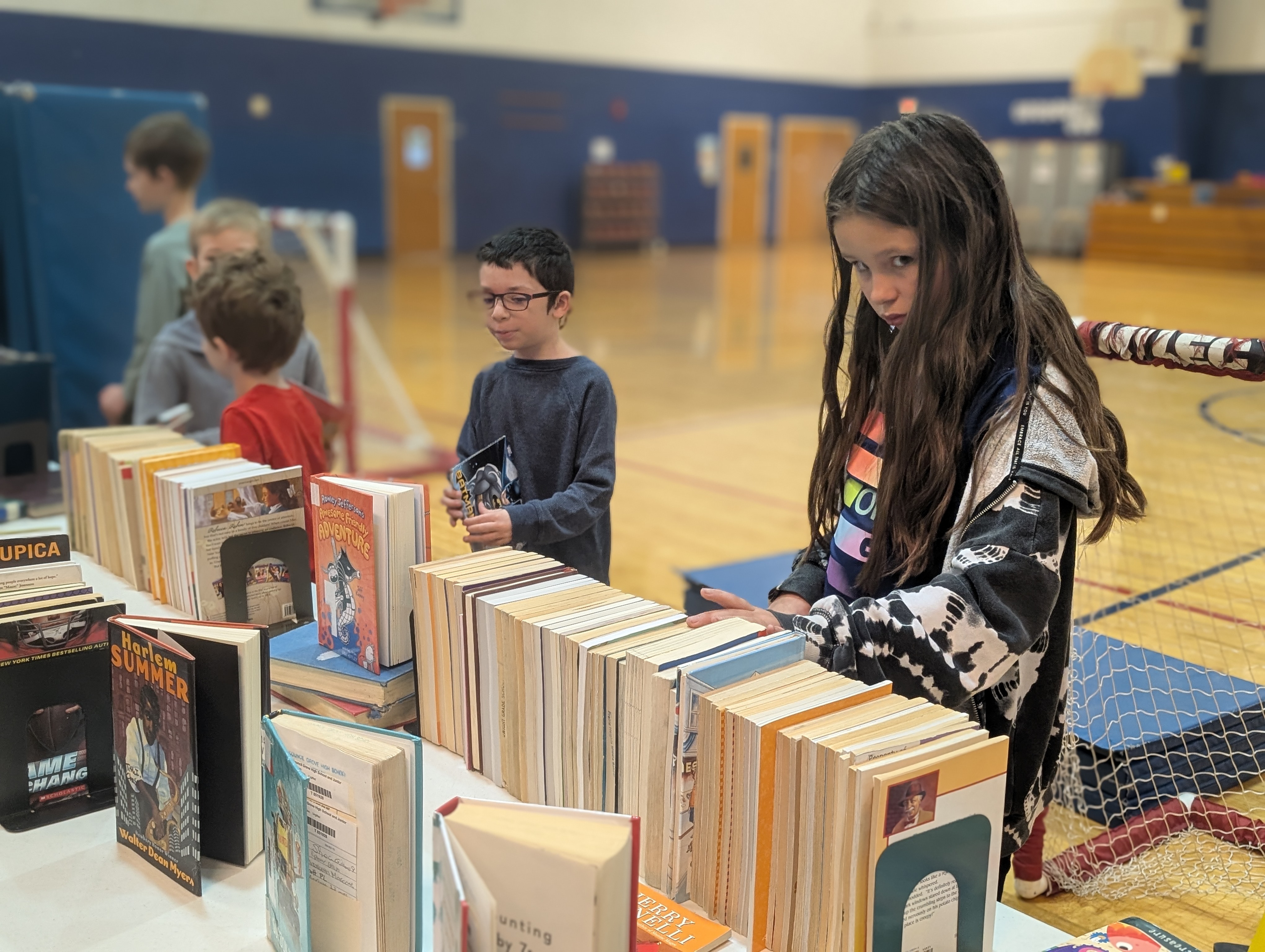 Children browse a table filled with books in a gymnasium, with a girl in the foreground looking towards the camera.