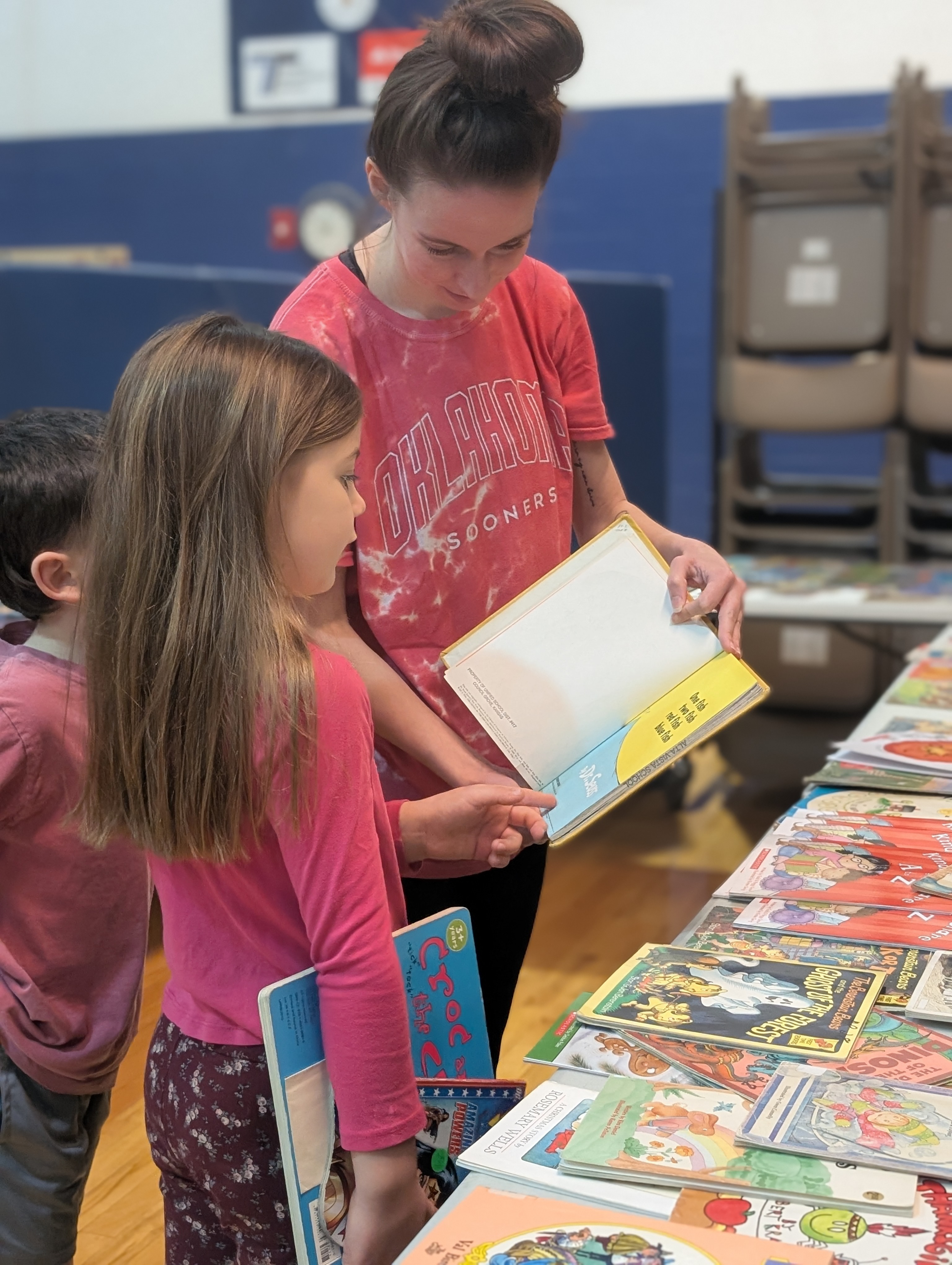 A woman in a red tie-dye shirt shows a book to two children at a table filled with books.
