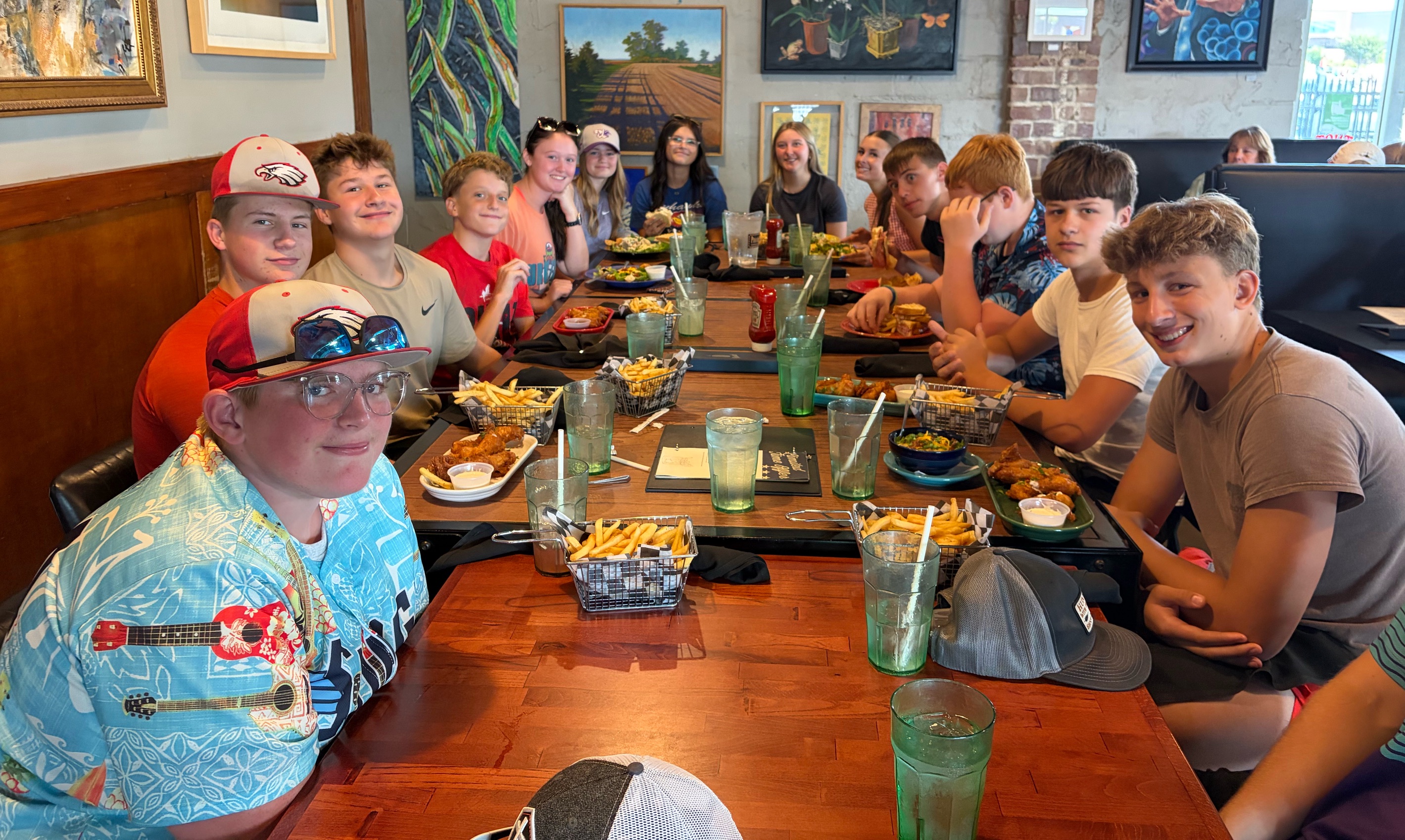 A group of young people, mostly teenage boys, sit at a wooden table in a restaurant, with plates of food and drinks in front of them.
