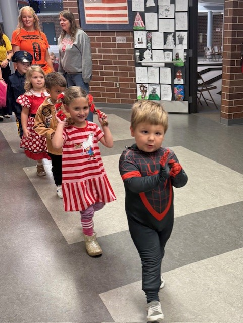 A line of young children in costumes walks down a school hallway, supervised by adults.