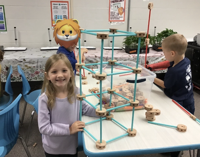 A smiling girl stands next to a large geometric structure built with blue rods and wooden connectors in a classroom.