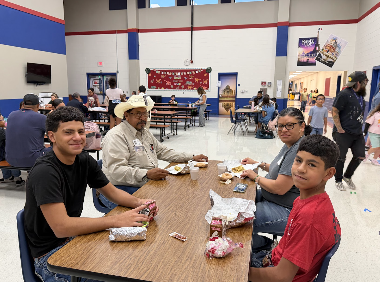 A family of four sits together at a wooden table in a school cafeteria, smiling as they enjoy a meal.