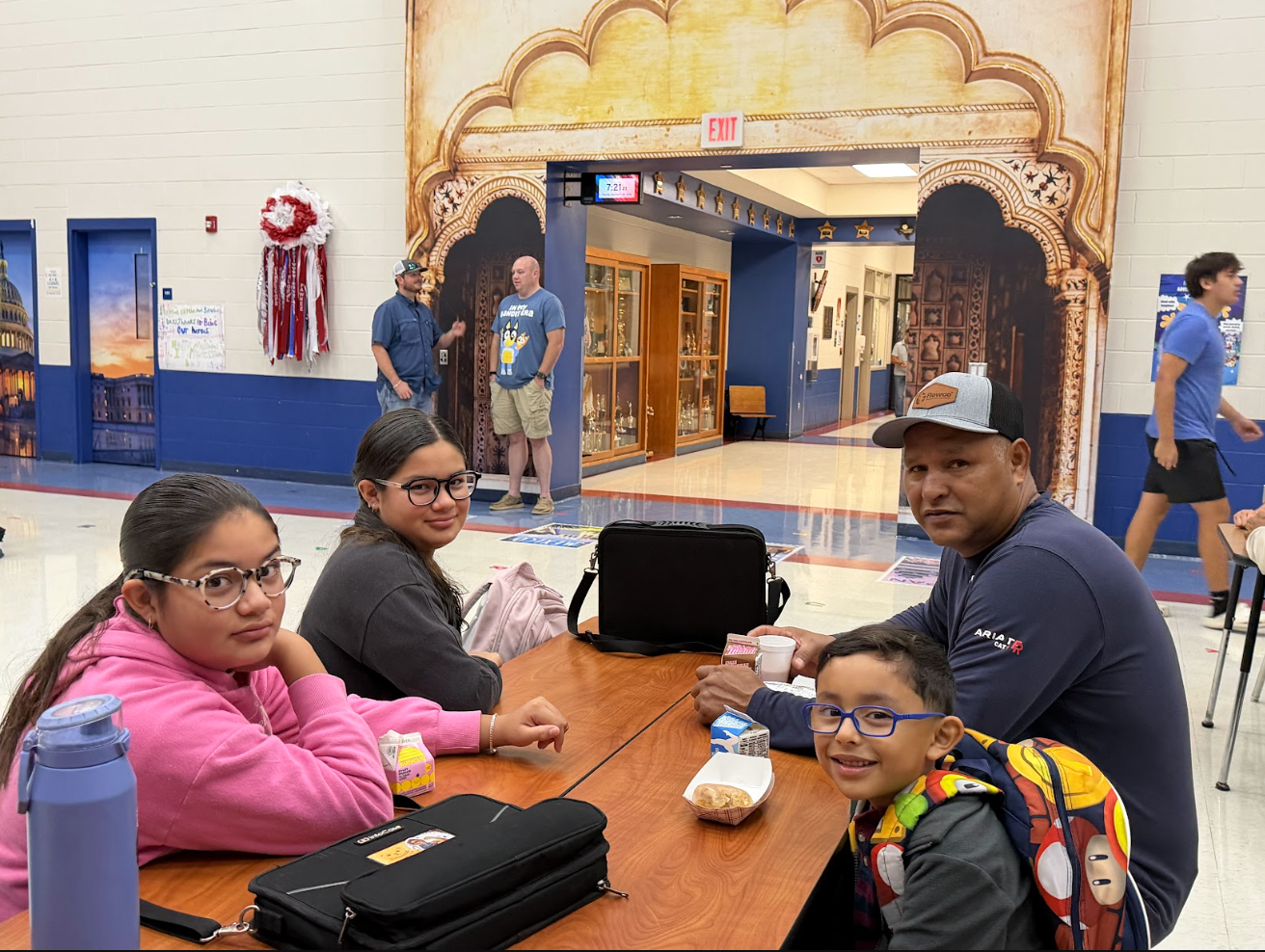 A family of four sits together at a cafeteria table, smiling for a group photo while eating lunch in a brightly lit school hallway.