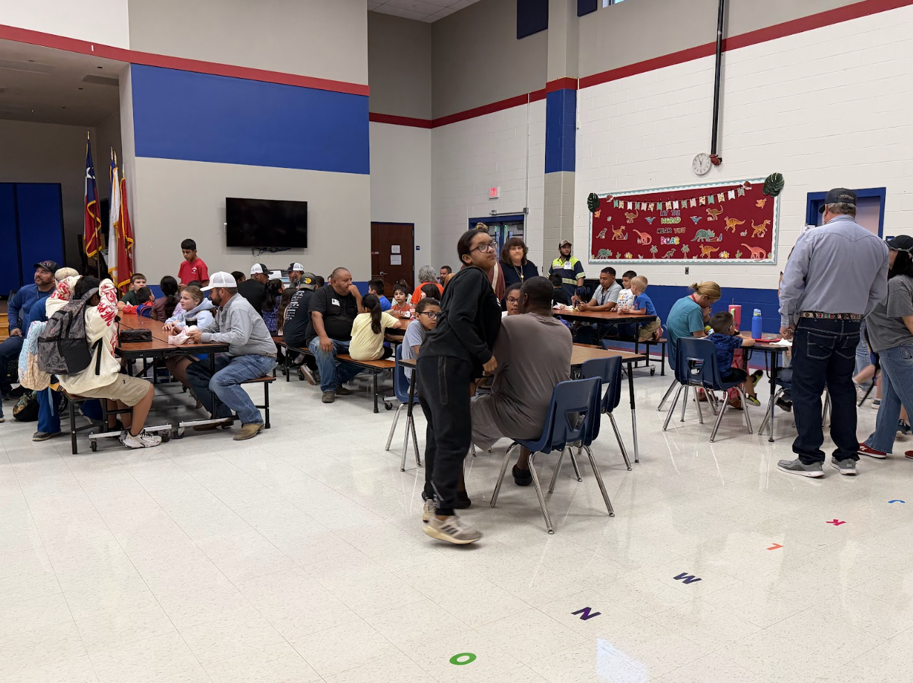 A bustling school cafeteria with adults and children seated at tables, socializing during an event.