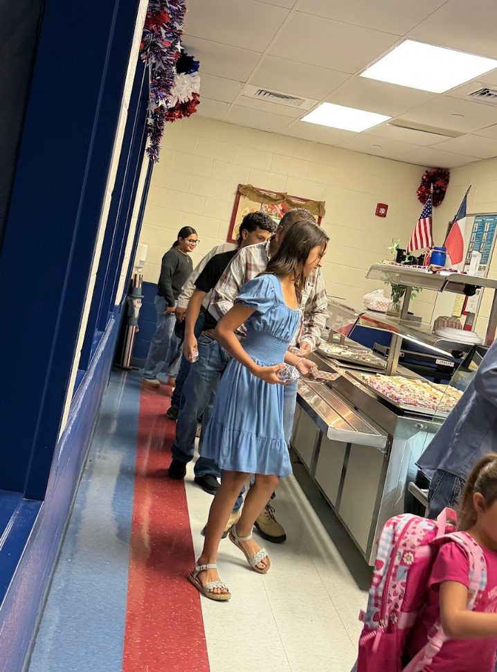 People line up in a school cafeteria to select food from serving counters.