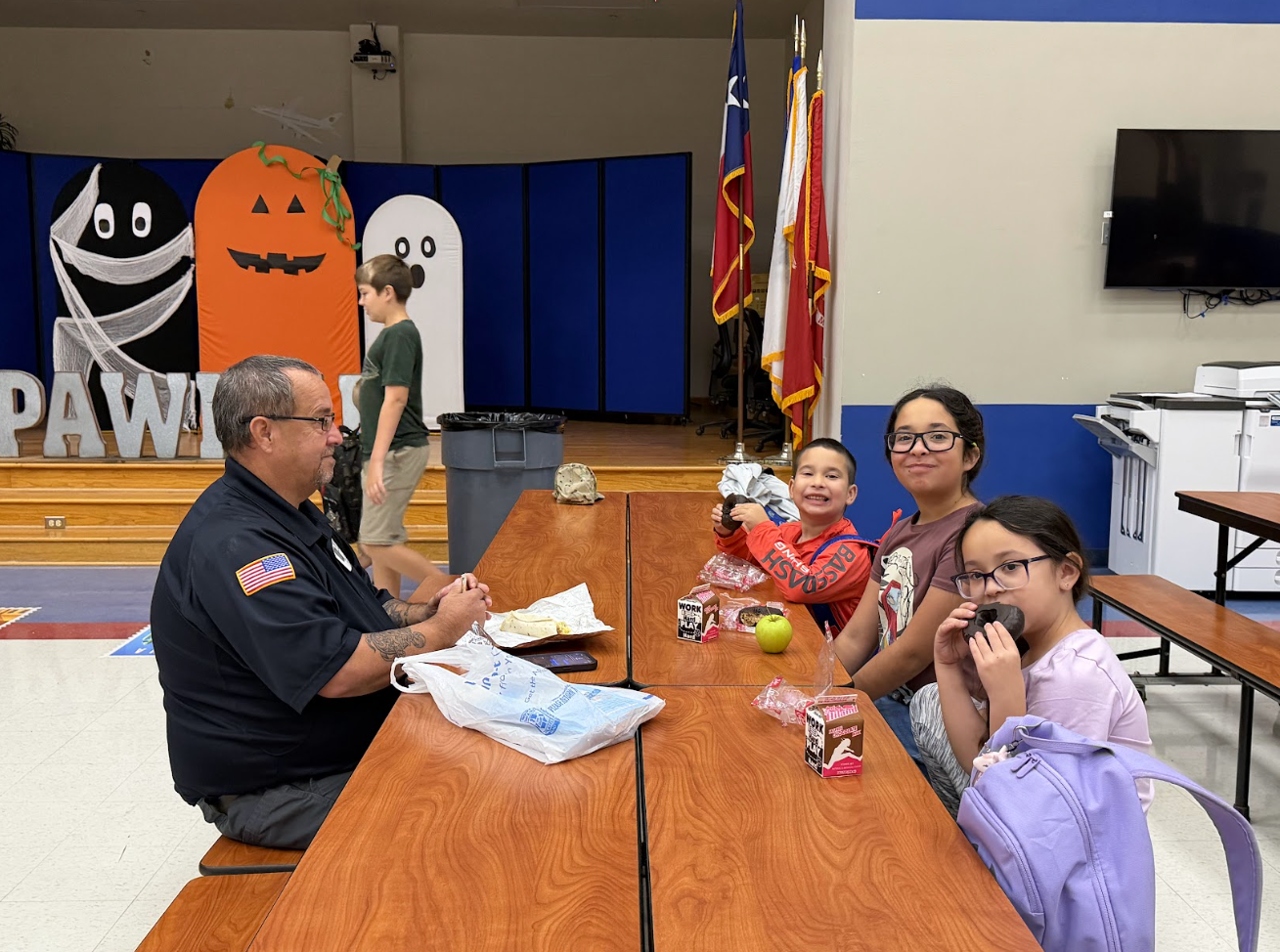 A school employee and students sit at wooden cafeteria tables, eating snacks and interacting in a decorated gymnasium.