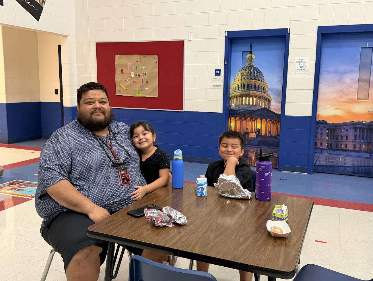 A smiling adult and young child sit next to an older child at a cafeteria table with lunch items.