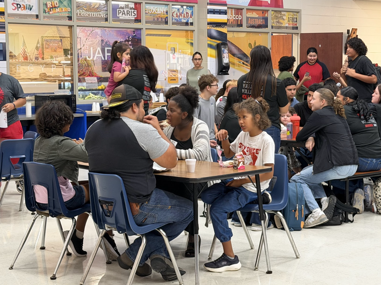 Students and families gather inside a school cafeteria, seated at tables and eating together.