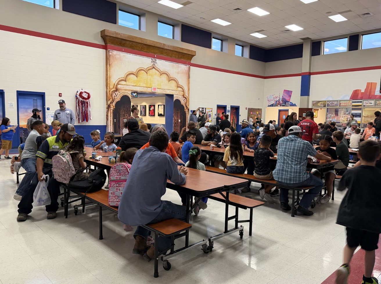A bustling school cafeteria filled with children and adults sitting at long tables, eating and socializing.