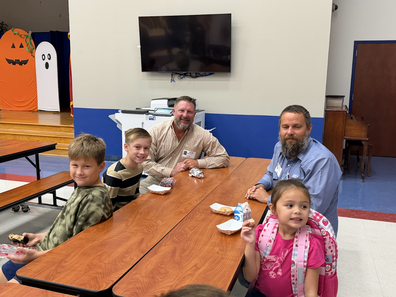 Two adults and three children sit together at connected wooden cafeteria tables, smiling at the camera.