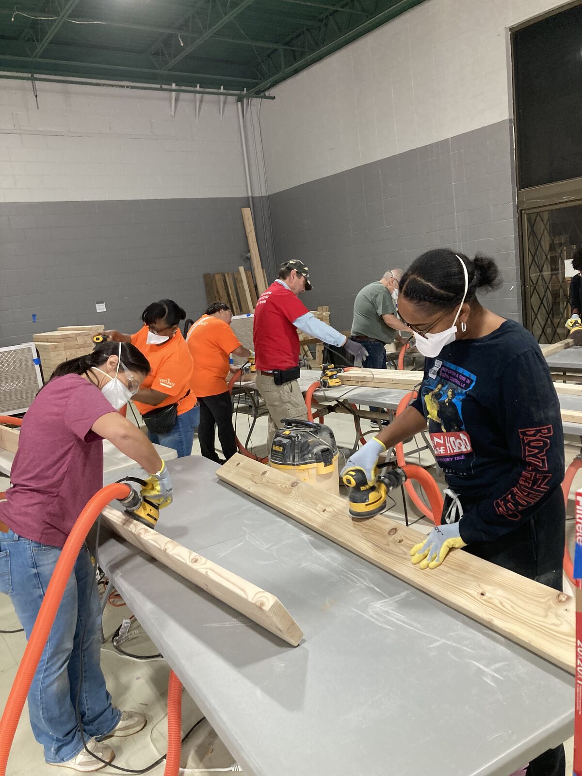 Group of volunteers wearing face masks and safety gear sanding wooden lumber at workbenches.
