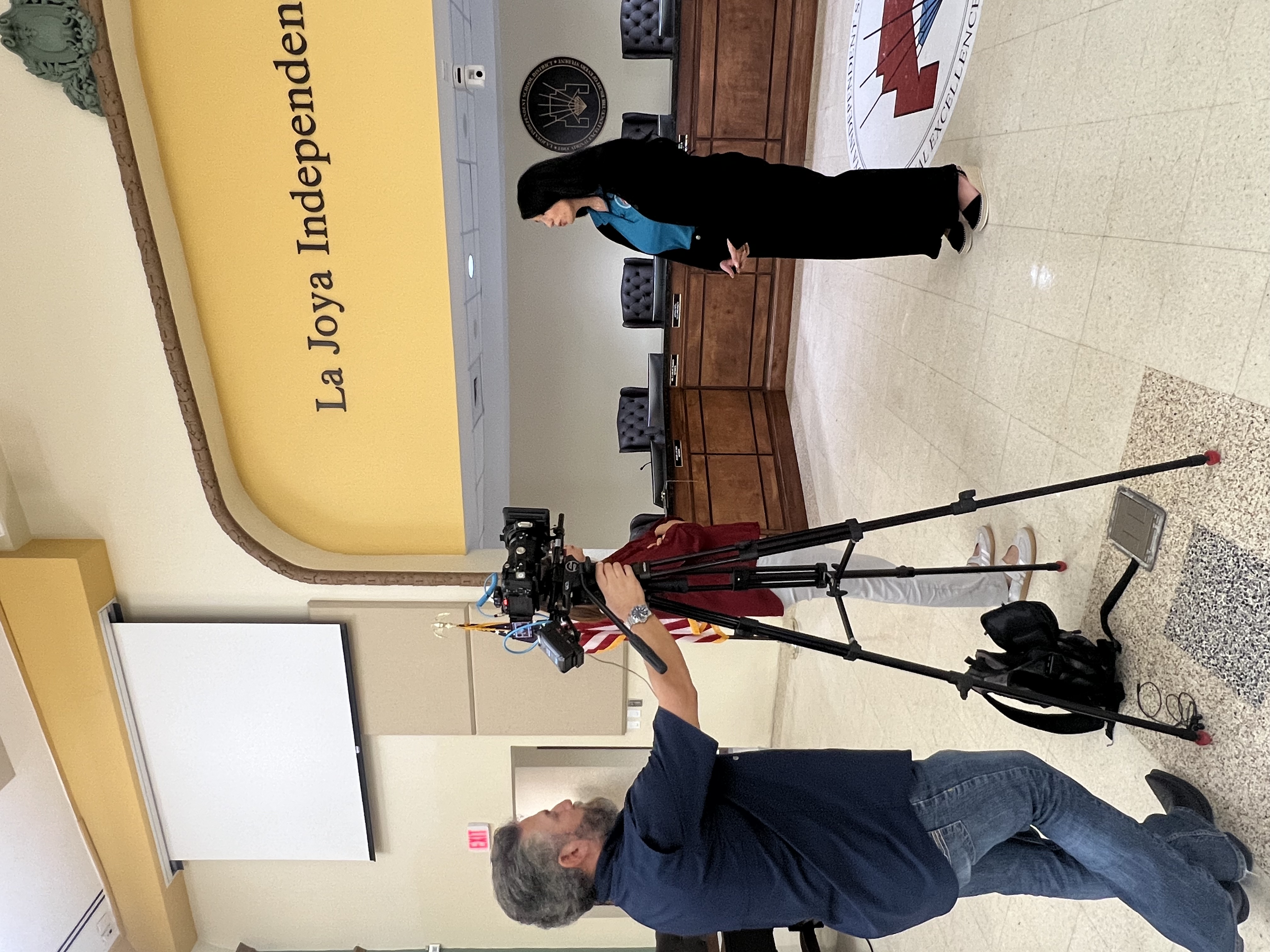 A person operates a video camera on a tripod, filming a woman in a black robe and blue shirt.