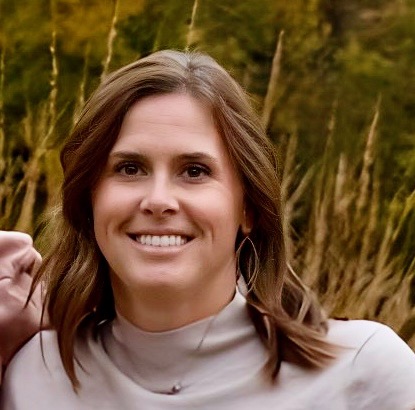 A woman with shoulder-length brown hair smiles warmly, wearing a light-colored turtleneck and a delicate necklace.