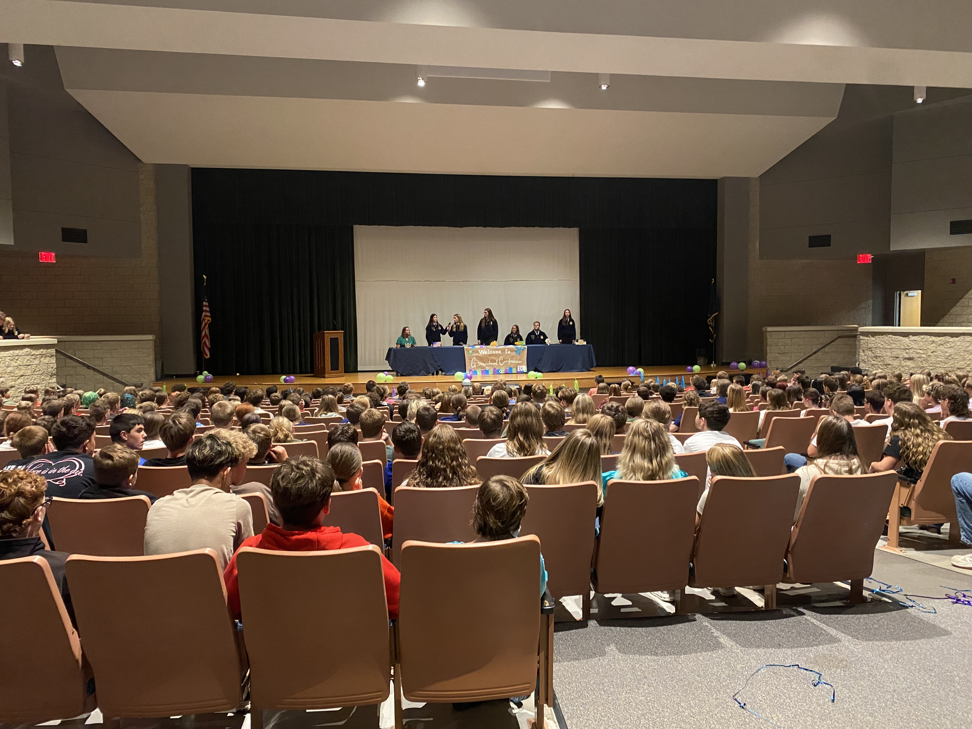 A large audience of students fills an auditorium, facing a stage where a panel of speakers is seated.