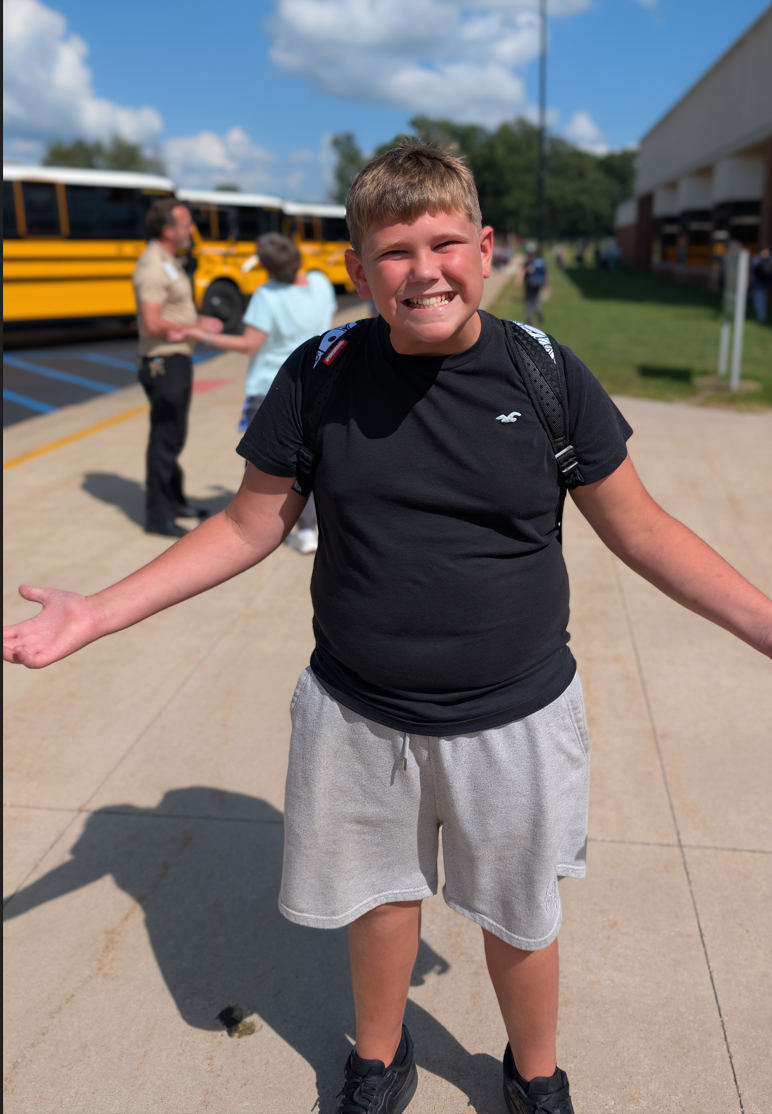 A smiling boy in a black t-shirt and grey shorts stands with arms outstretched in front of school buses.
