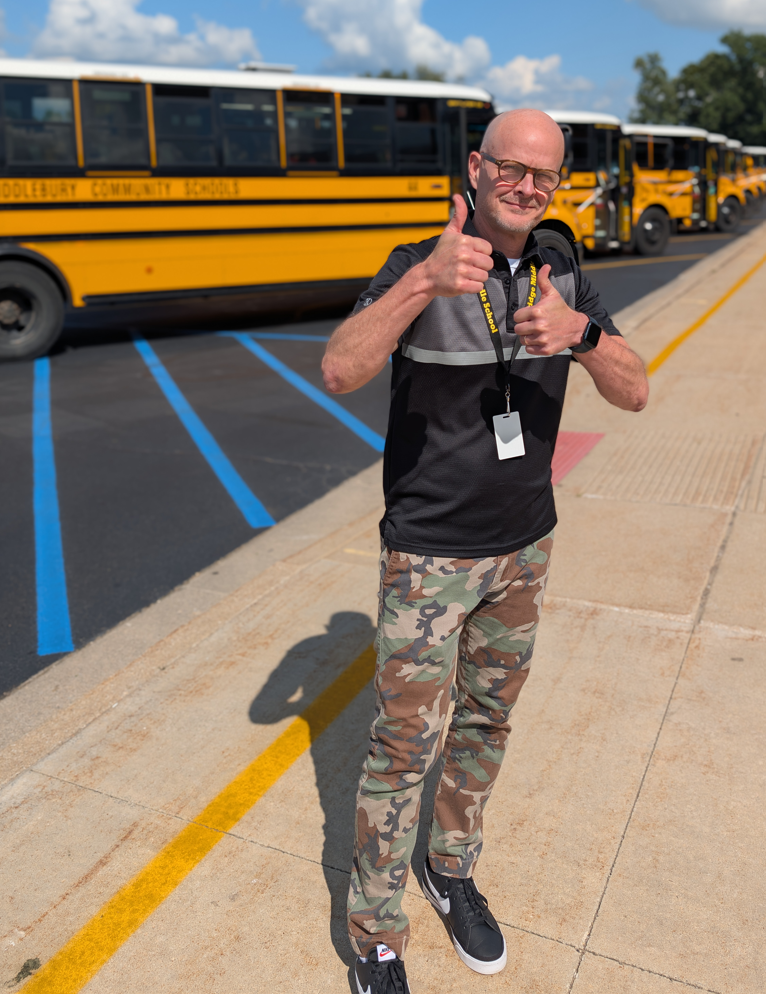 A man in camouflage pants and a black shirt gives two thumbs up in front of a line of yellow school buses.