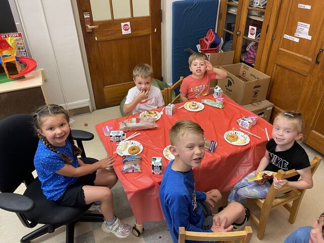 Children sit around a red-covered table with snacks and drinks, smiling at the camera.
