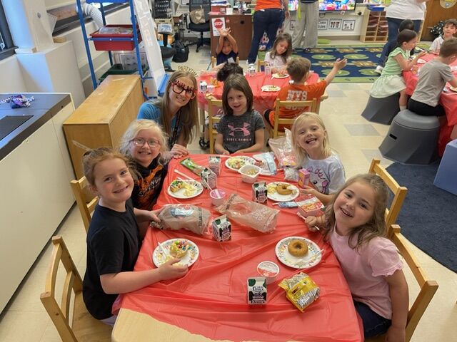 Children and adults sit around a table covered with a red tablecloth, enjoying snacks and drinks.