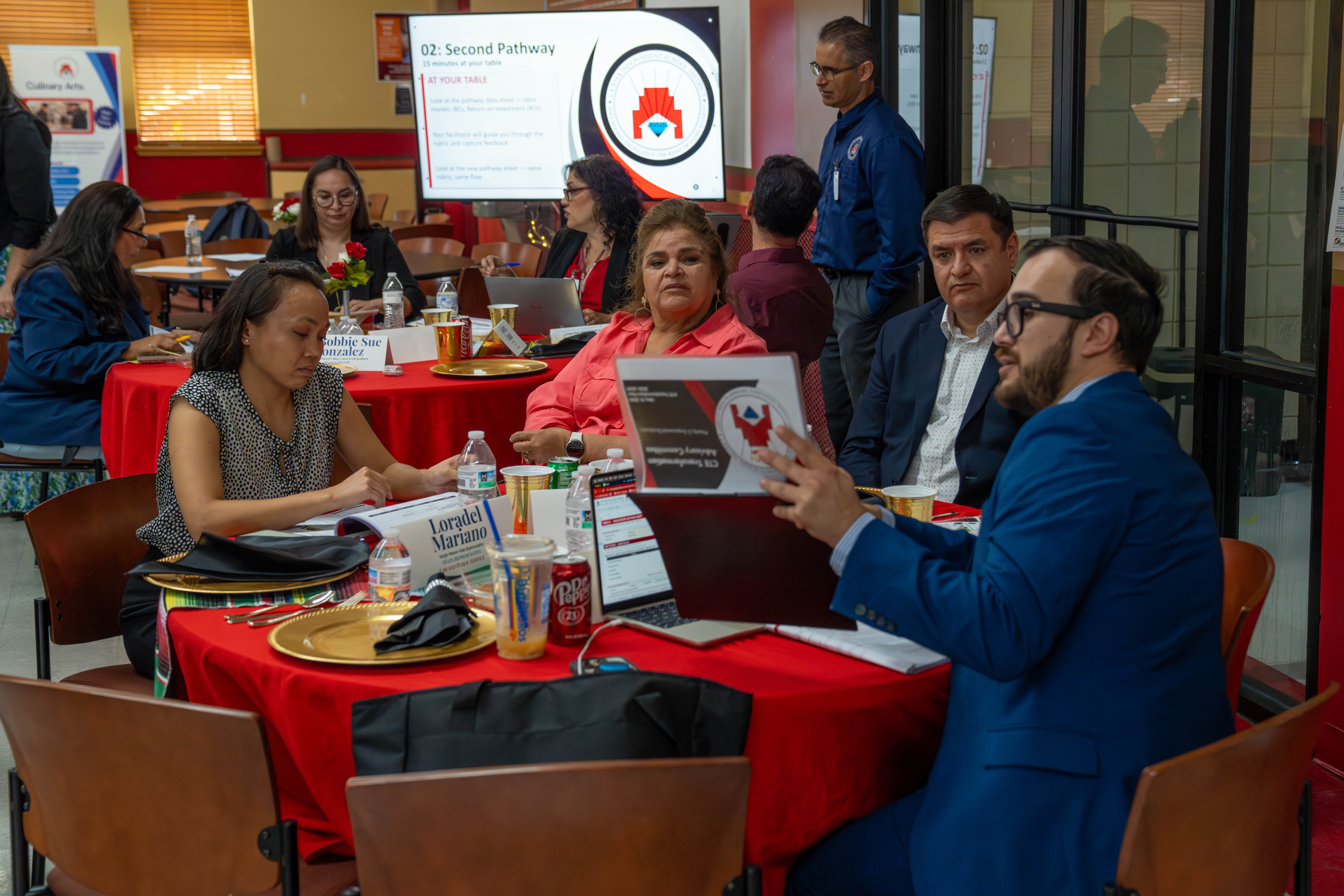A group of people are seated at round tables covered in red tablecloths, engaged in a presentation displayed on a large screen.
